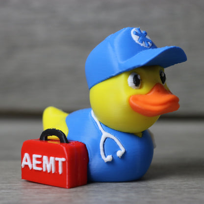 Rubber duck wearing a blue cap and holding a red suitcase with 'AEMT' on it, on a gray background.